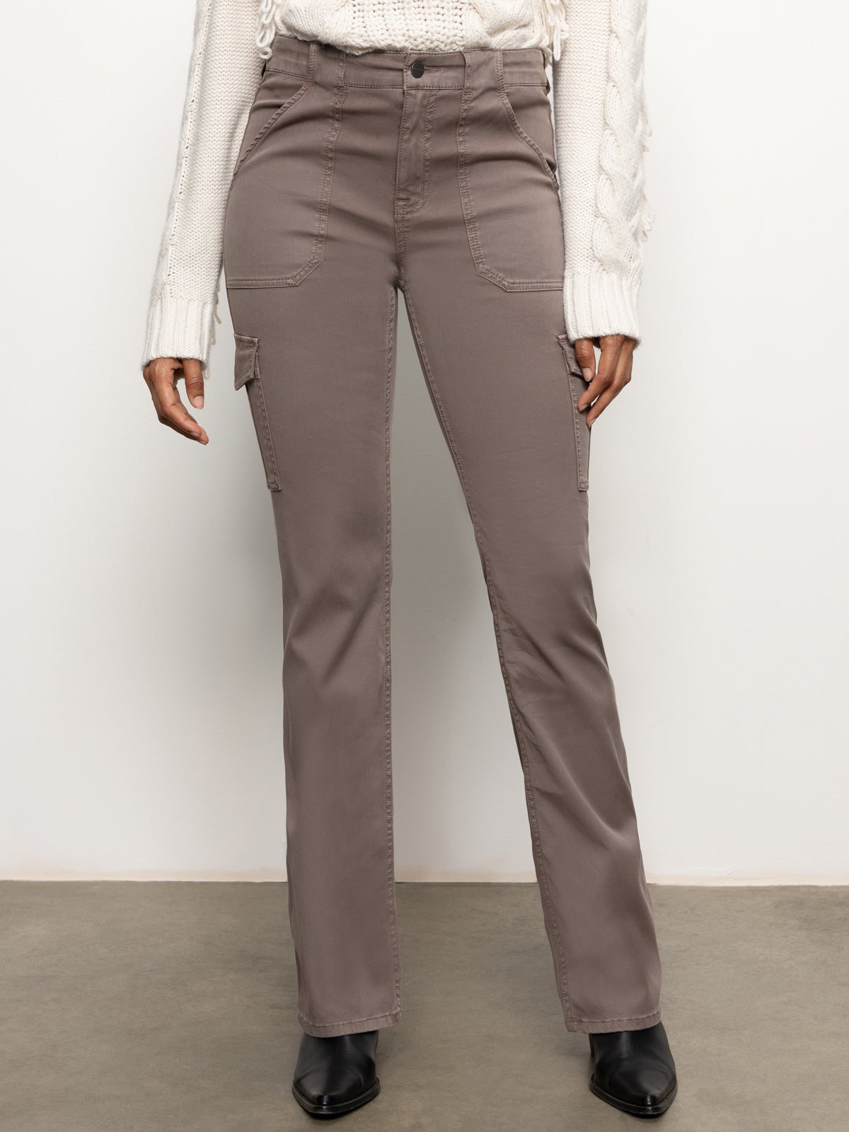 A person is wearing the Sculpted Hayden Bootcut Cocoa pants from Sanctuary Clothing, featuring a high-waisted fit with a front button and pockets. Pairing these stylish brown cargo pants with a white knit sweater and black ankle boots creates an elegant look in a neutral indoor setting.