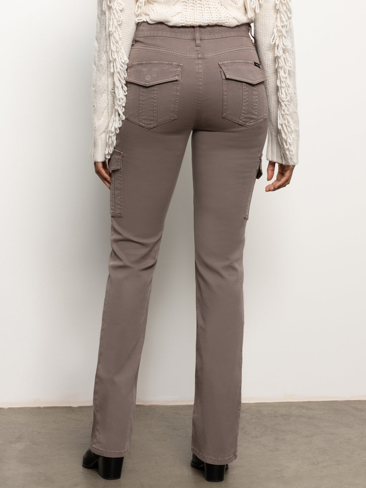 Rear view of a person wearing the Sculpted Hayden Bootcut Cocoa pants by Sanctuary Clothing. They stand on a light-colored floor against a plain background, dressed in a cream sweater with textured patterns and taupe cargo pants featuring pockets.