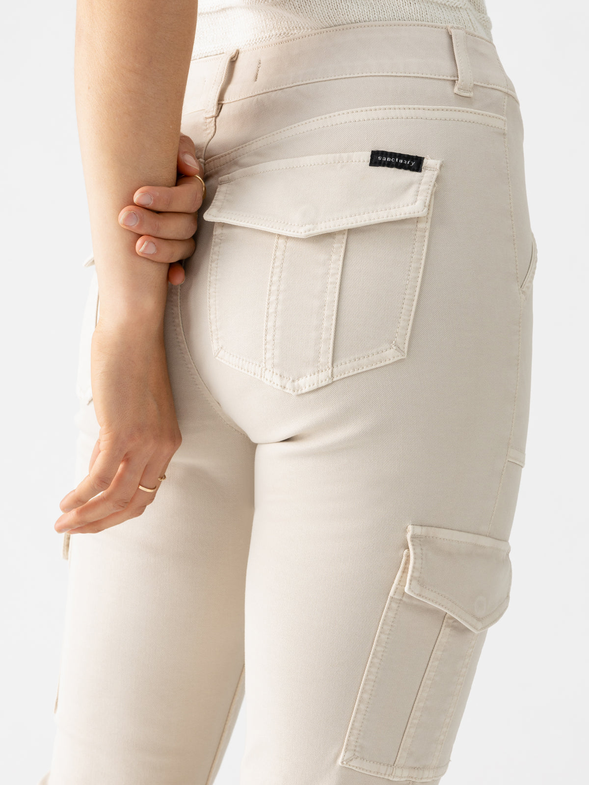 Close-up of a person standing with their hand resting on the back pocket. They are wearing Sanctuary Clothing's Sculpted Hayden Bootcut pants in Toasted Almond, featuring multiple pockets including a visible thigh pocket and a small black label on the back pocket. The person's upper body is partially visible.