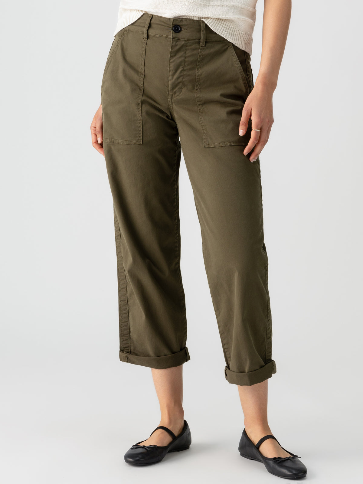 A person is standing against a plain background, wearing Sanctuary Clothing's Cruiser Chino Burnt Olive pants with large front pockets and a white top. They have their hands resting casually near the pocket areas. They are also wearing black flats.