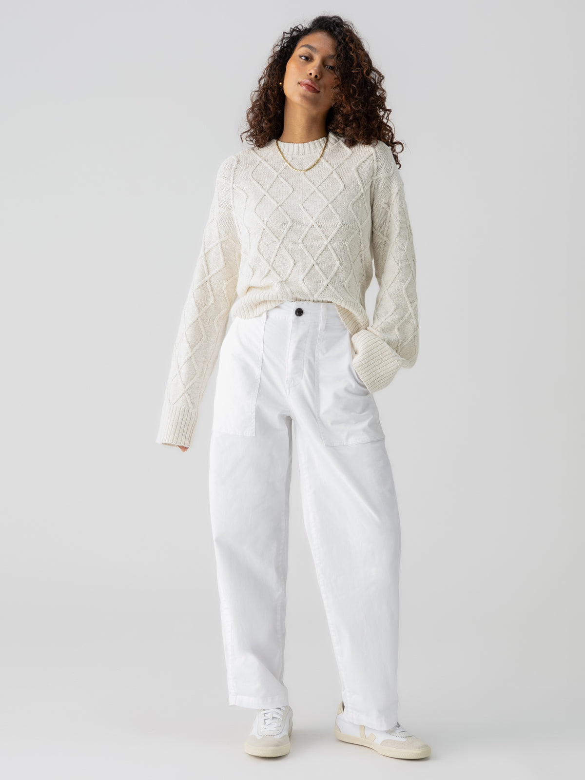 A woman with curly hair stands against a plain background, confidently serene. She is dressed in a white cable-knit sweater, Cruiser Chino Chalk high-waisted pants by Sanctuary Clothing, and white sneakers. Her right hand hangs relaxed by her side.