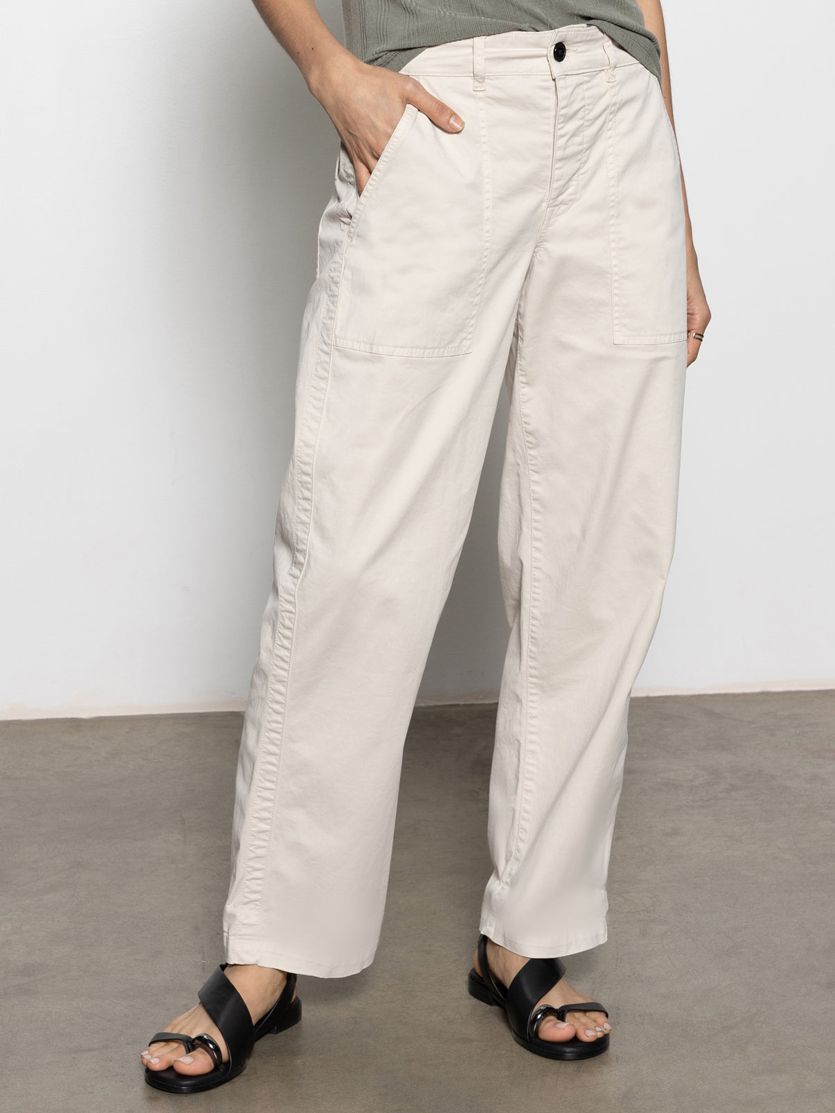 A person wearing Sanctuary Clothing's cruiser chino light oat pants and black sandals stands on a gray floor with one hand in a pocket. The upper body is not visible, set against a plain white wall.