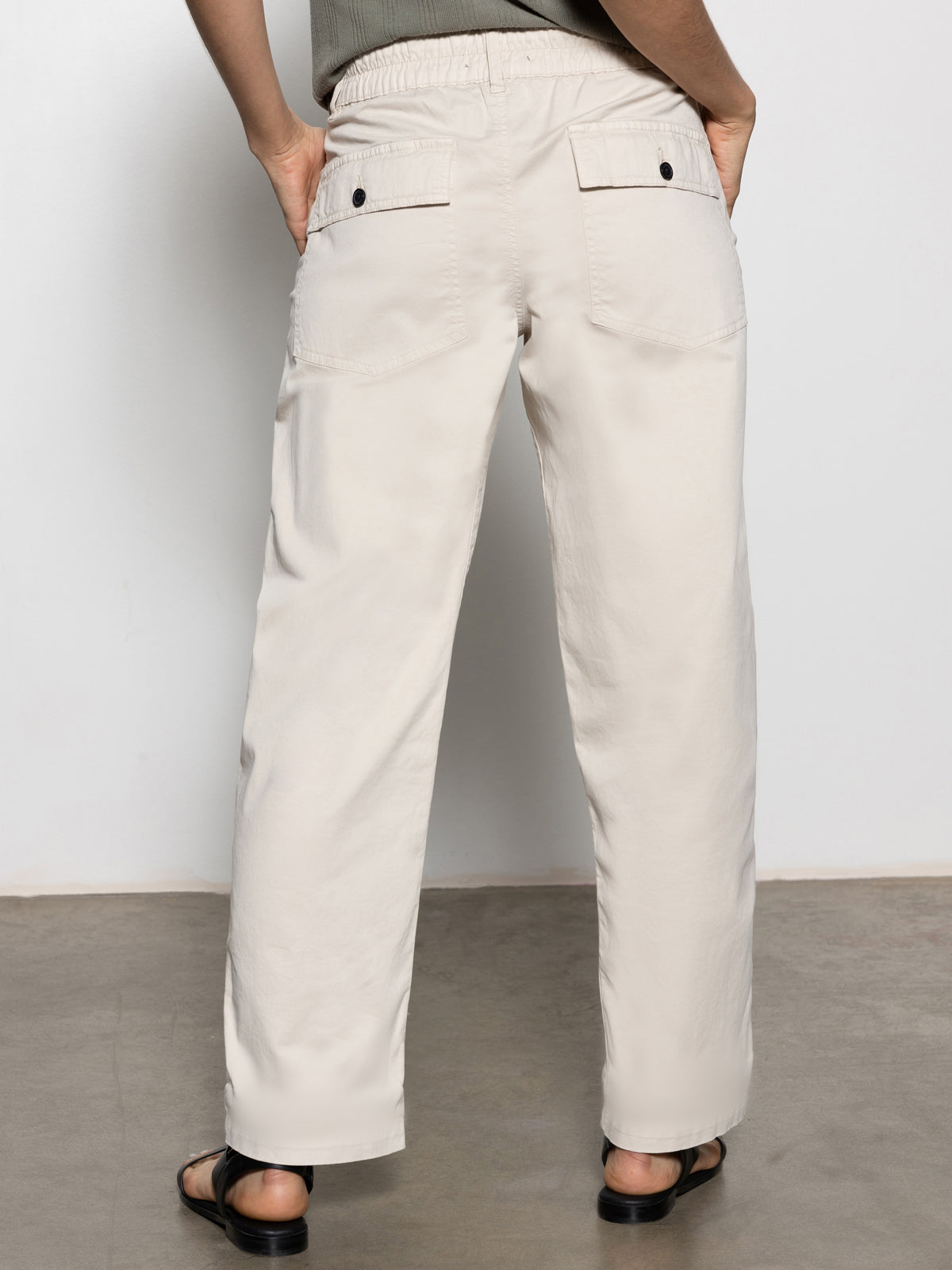 A person is wearing Sanctuary Clothing's Cruiser Chino Light Oat pants featuring buttoned back pockets, paired with a dark green top and black sandals, standing against a plain light background.