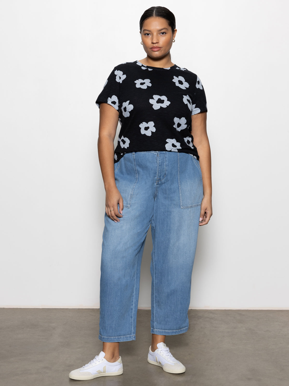 A person stands against a plain backdrop, wearing Sanctuary Clothing's black t-shirt adorned with white flower designs and cruiser chino wide-leg pants from the Waverly Inclusive Collection. They have short dark hair, wear white sneakers, and look confidently at the camera.