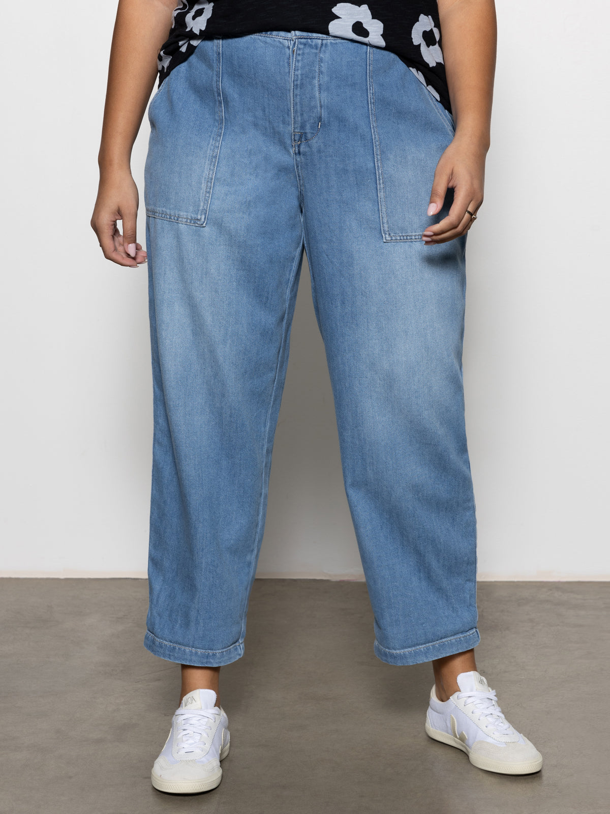 A person in light blue denim jeans and white sneakers stands on a concrete floor, wearing Sanctuary Clothing's Cruiser Chino from the Waverly Inclusive Collection. They are also sporting a black top with white floral patterns, and the image is cropped at the torso.