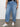 A person wearing the Cruiser Chino Waverly from the Inclusive Collection by Sanctuary Clothing, light blue with large front pockets, and a black floral top is standing on a concrete floor. Their feet are in white sneakers. The background is a plain off-white wall.