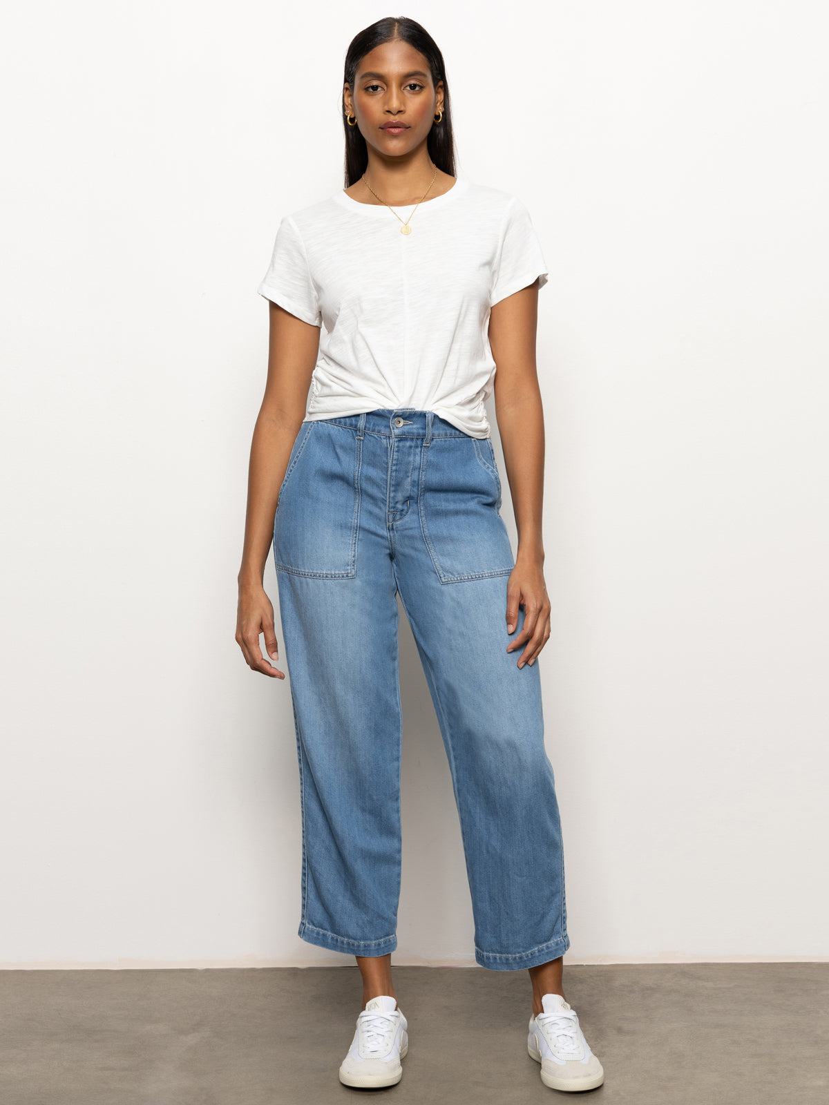 A person stands against a plain backdrop, dressed in the Cruiser Chino Waverly by Sanctuary Clothing. They pair it with a white T-shirt, high-waisted blue jeans, and white sneakers. Sporting long hair and a gold necklace, they maintain a relaxed, front-facing pose.