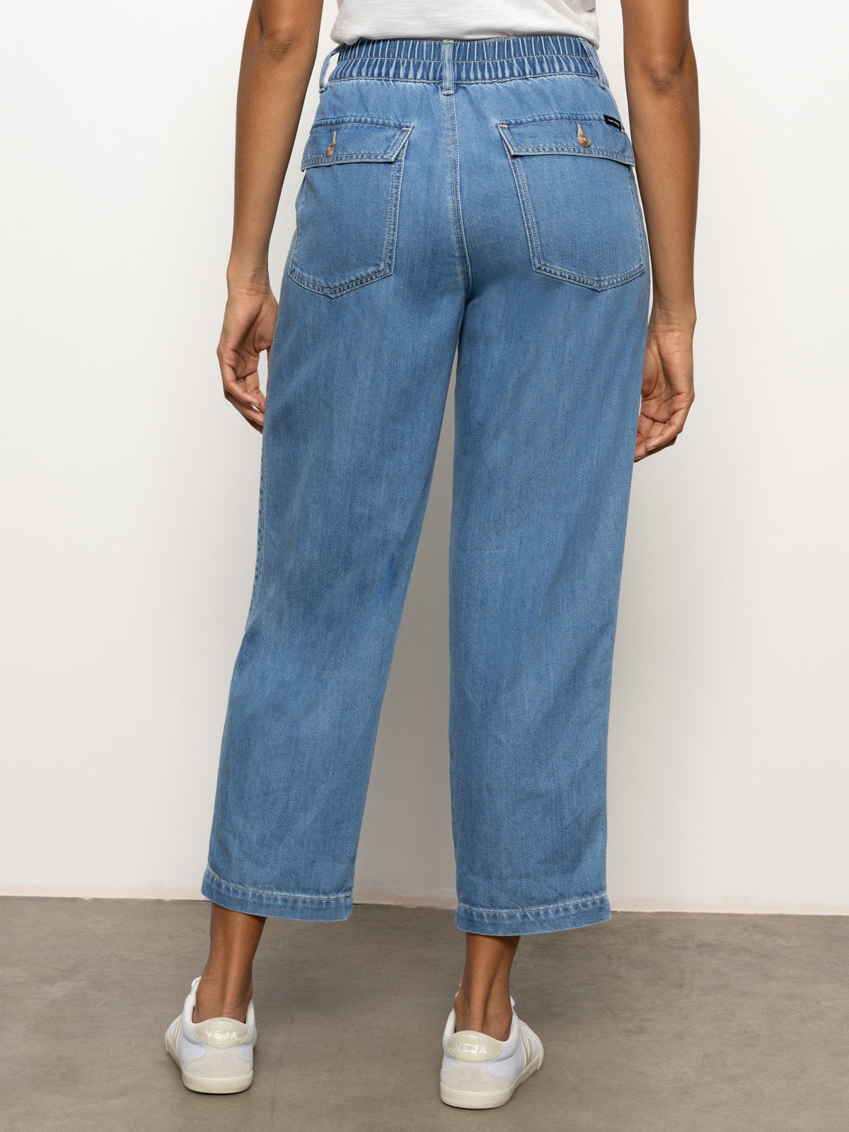 A person is seen from the back wearing the Sanctuary Clothing's cruiser chino waverly: relaxed-fit, high-waisted blue jeans featuring a cinched elastic waistband and two back pockets. They pair it with white sneakers against a plain light-colored background.