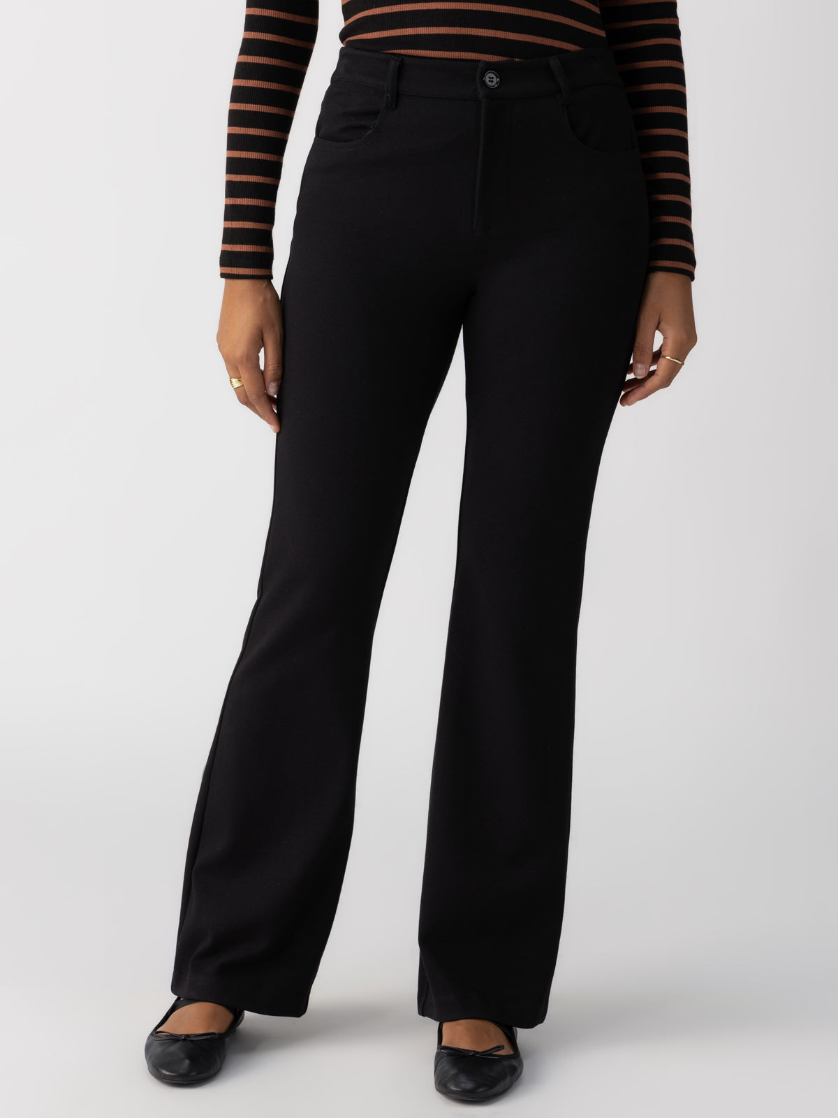 A person is wearing Sanctuary Clothing's Maya Flare Black pants, paired with a black and brown striped long sleeve shirt. Their footwear consists of black flat shoes. The image does not show the person's face, focusing only on the torso and lower body against a plain background.