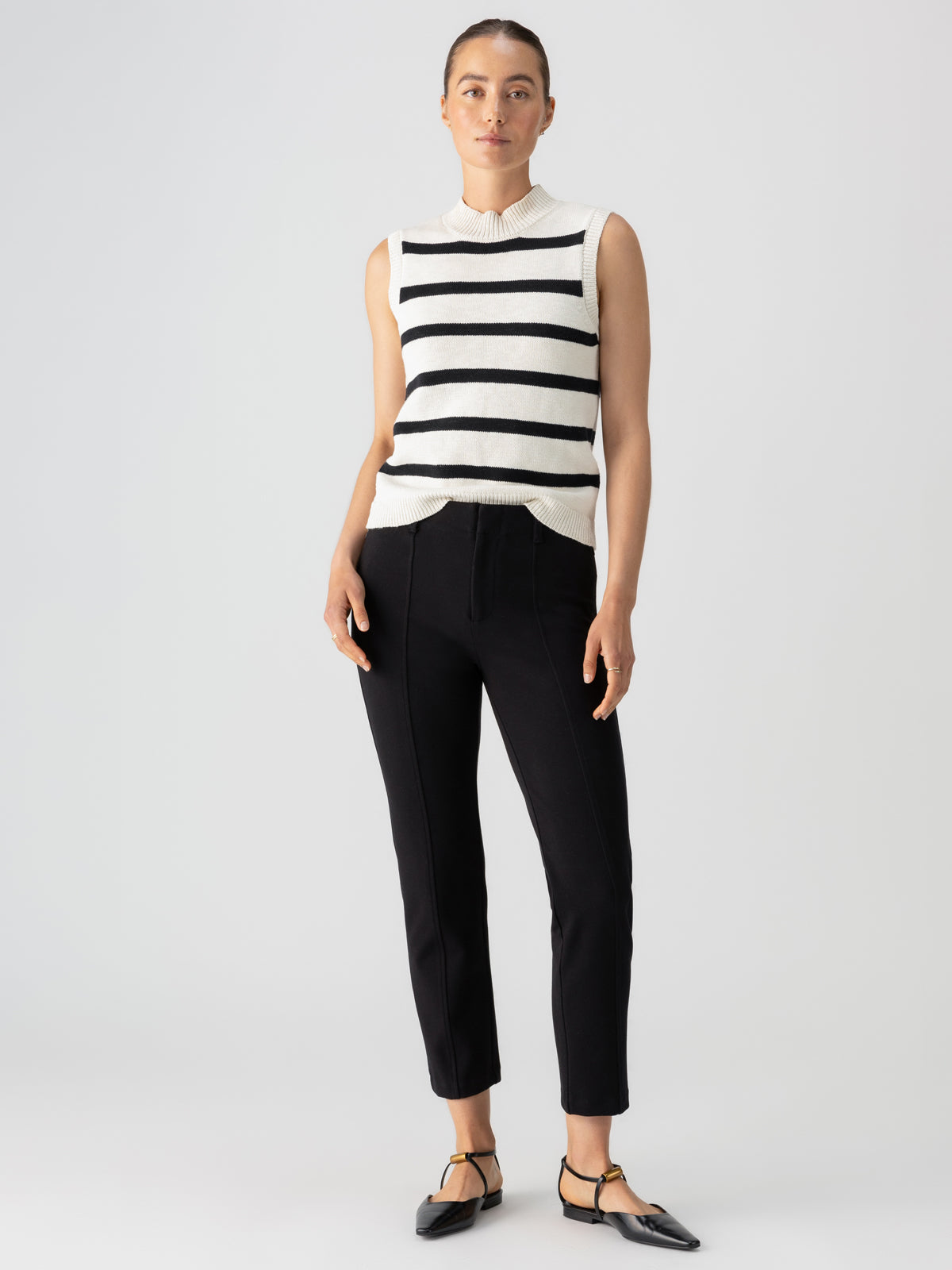 A person stands against a neutral background, wearing a sleeveless white sweater with black horizontal stripes, paired with the Sanctuary Clothing Cassie Legging Black and black flats featuring a pointed toe and gold strap detail. The individual has their hair pulled back.