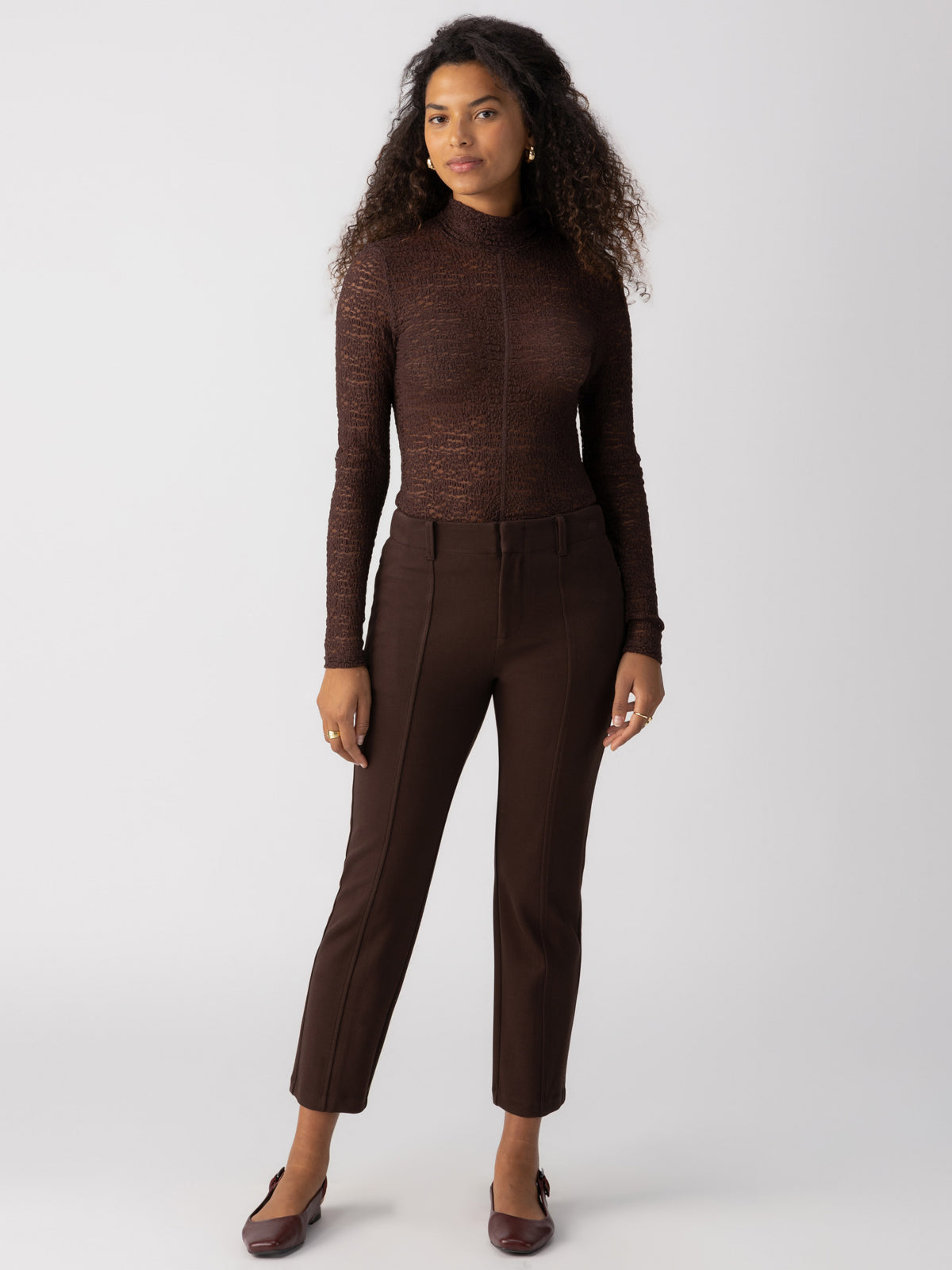 A person with long, curly hair stands against a plain background, wearing Sanctuary Clothing's Cassie Legging in Coffee along with a coordinated long-sleeve, high-neck brown top and brown shoes. They also sport minimal jewelry and face the camera with a neutral expression.