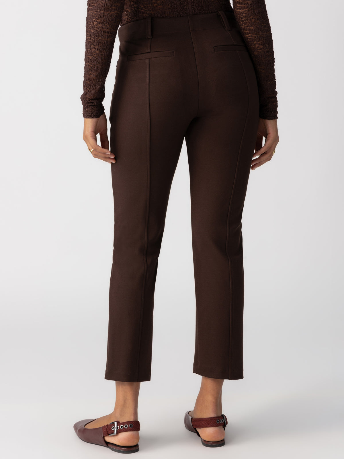 A rear view of a person standing, wearing Sanctuary Clothing's "Cassie Legging Coffee," which are high-waisted brown pants featuring seam details and flat pockets. They are also donning a textured brown long-sleeve top and burgundy flats with ankle straps. The background is plain white.