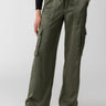 A person is wearing the "Satin Cargo Wide Leg Dark Olive" pants by Sanctuary Clothing, which feature multiple pockets and a drawstring waist. They have paired this outfit with black ballet flats and a black top. The background is plain and gray, and their left hand rests by their side.