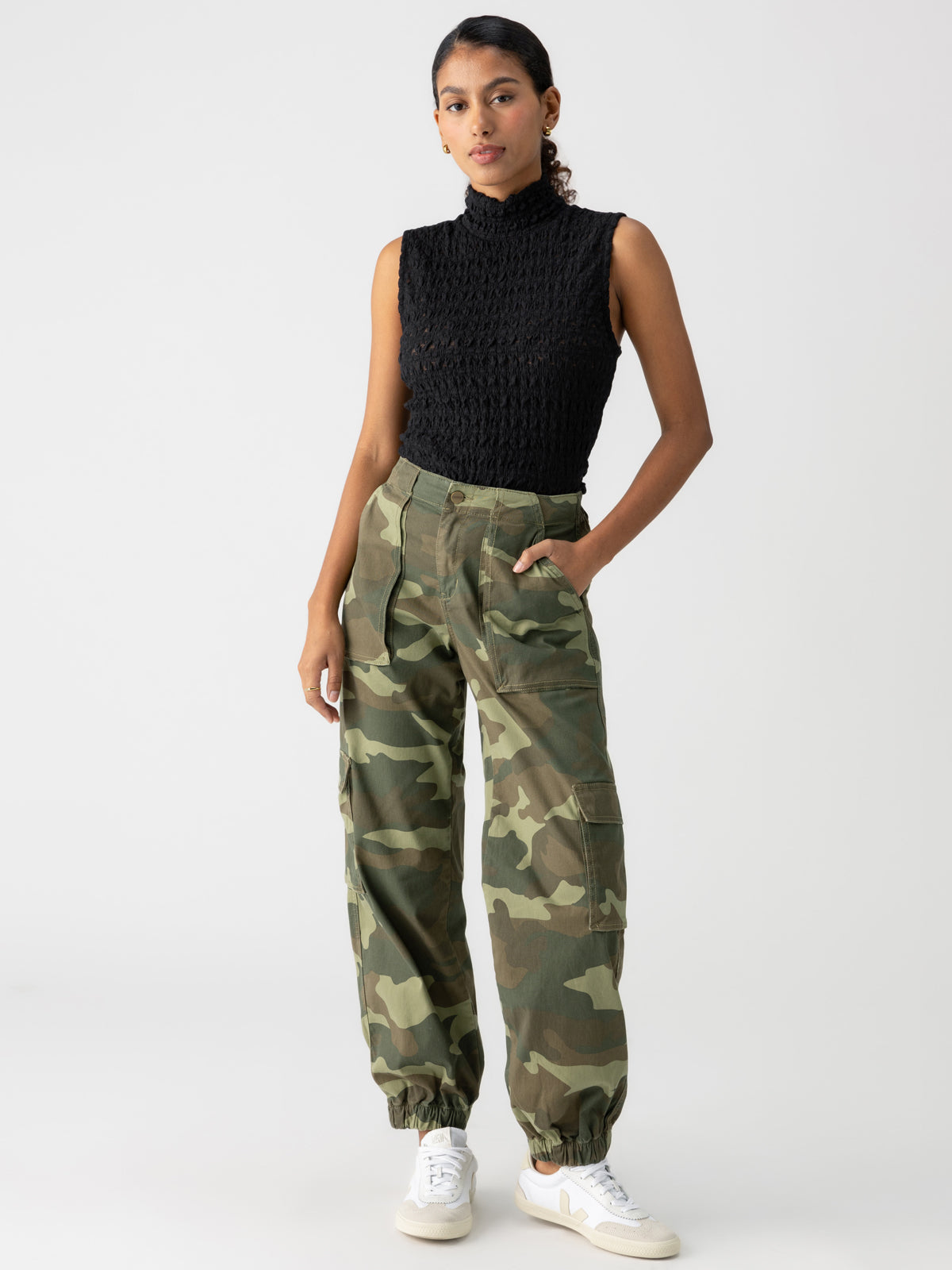 A person with tied-back hair wears the Cyber Cargo Renew Camo pants from Sanctuary Clothing, paired with a sleeveless black textured top, standing in a confident pose with one hand in their pocket. They are also wearing white sneakers against a plain white background.