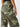 The person is wearing the Cyber Cargo Renew Camo pants from Sanctuary Clothing. The high-waisted, green camouflage pants feature multiple pockets and are paired with a black waistband from another garment. Their right arm rests partially visible at their side against a plain, light-colored background.