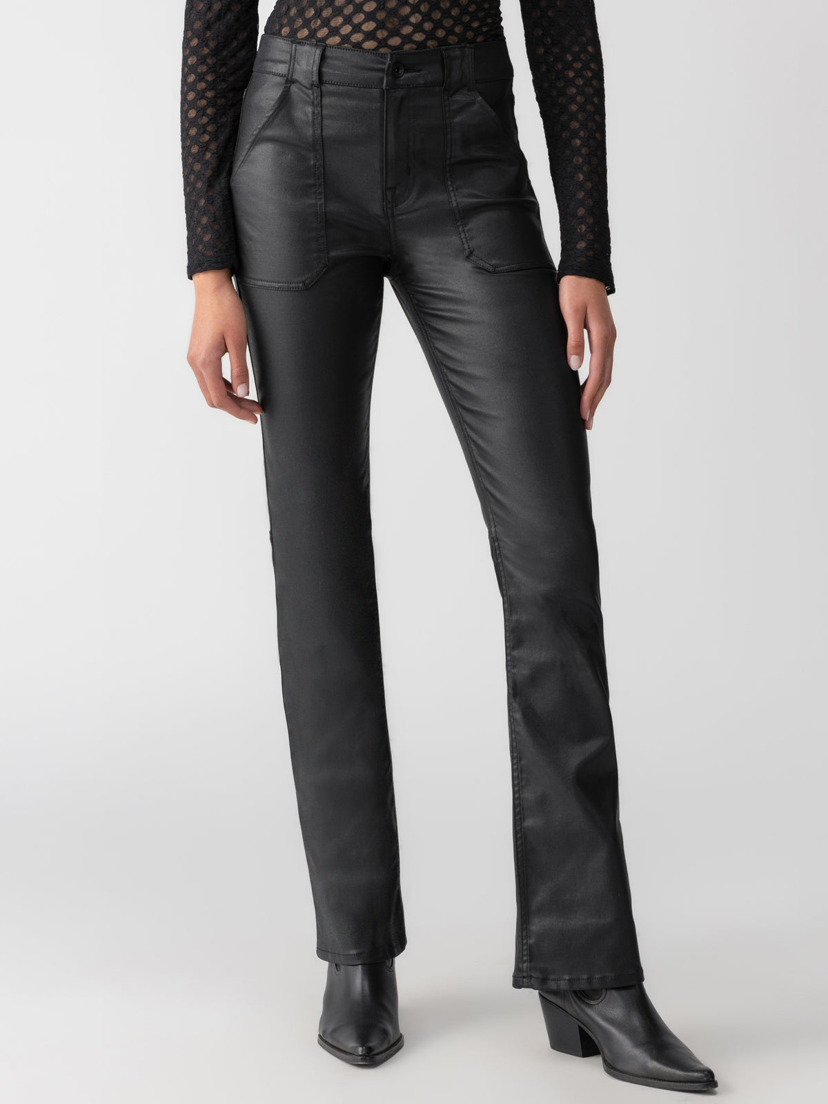 A person is standing, wearing the Sanctuary Clothing Slick Hayden Bootcut Pant in black paired with a black textured long-sleeve top. They also have on black ankle boots with a slight heel. The background is plain and minimalistic.