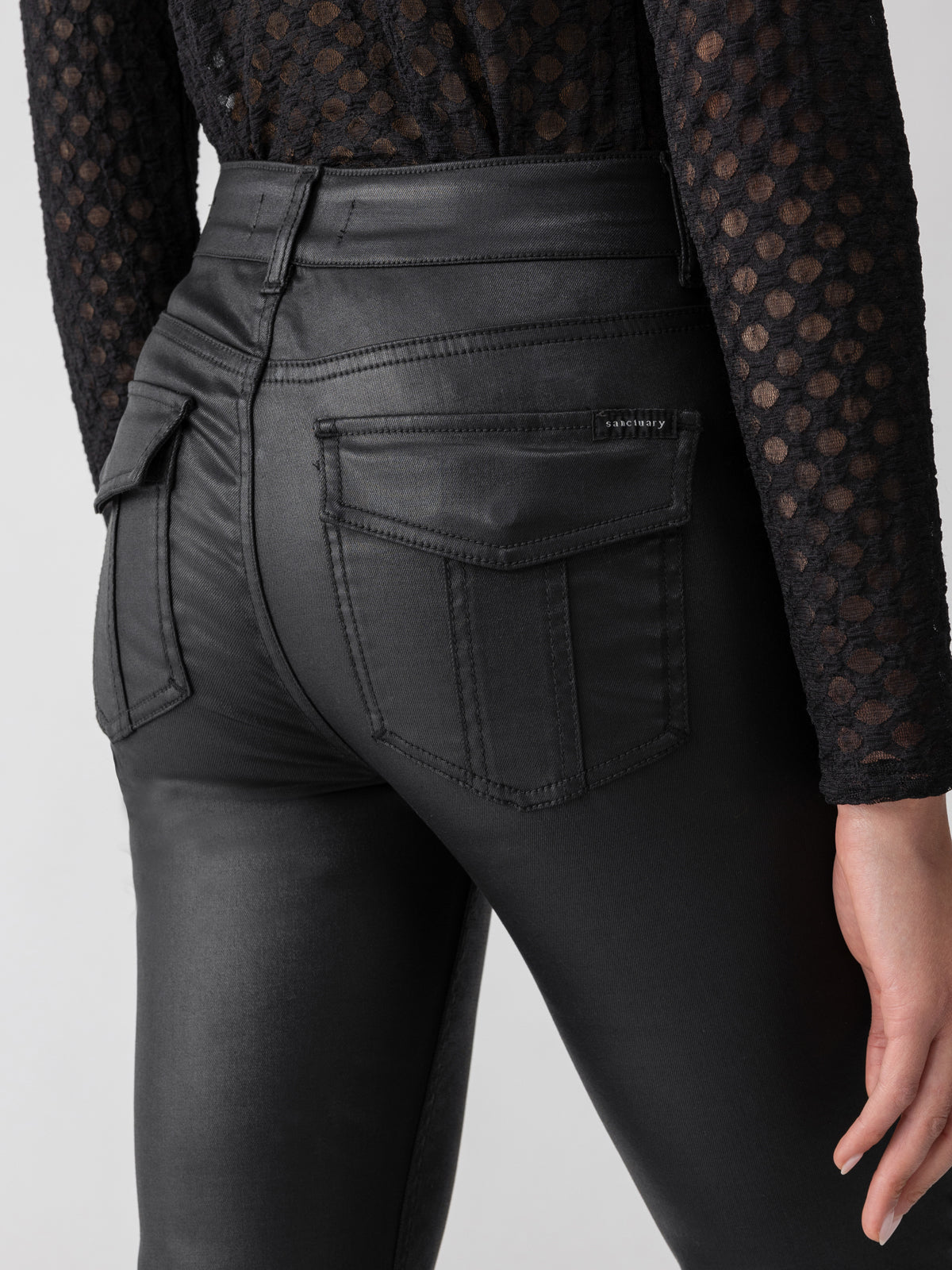 A person is shown from the waist down wearing the high-waisted, tailored-fit Sanctuary Clothing Slick Hayden Bootcut Pant Black, featuring visible stitching and back pockets. The person is also sporting a black, long-sleeve, semi-transparent mesh-textured shirt.