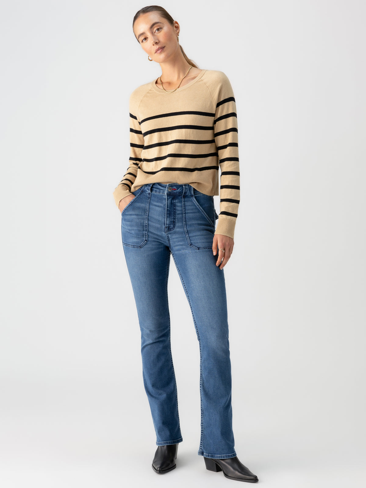 A person wearing the Sculpted Hayden Bootcut Lucky Streak by Sanctuary Clothing, paired with a beige sweater featuring black horizontal stripes, is standing against a plain white background. They are donning blue jeans and black heeled boots, with their right hand in their pocket and their left hand hanging by their side.