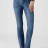 A person wears Sanctuary Clothing's Sculpted Hayden Bootcut Lucky Streak jeans, which feature front pockets and visible stitching. They pair the high-waisted blue jeans with a beige and black striped sweater, completing the look with black pointed-toe shoes. The background is light gray.