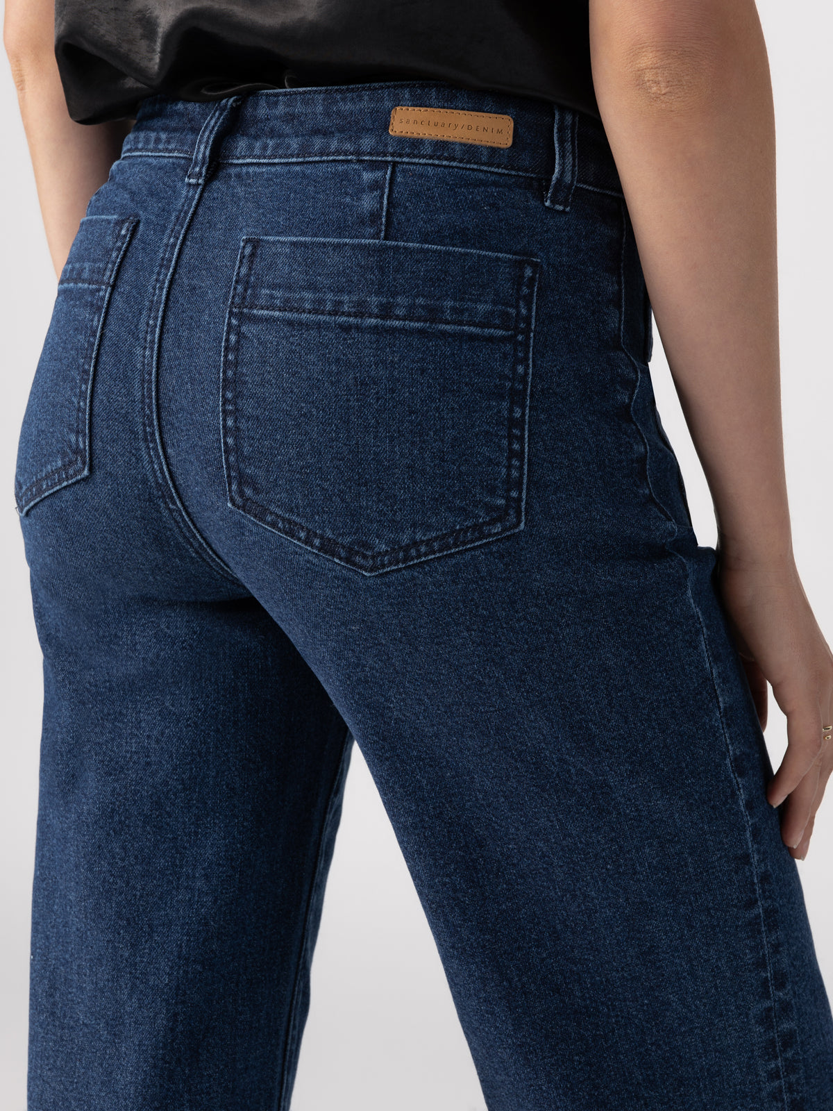 Close-up of a person wearing Sanctuary Clothing's Marine Full Length Aspen jeans. The focus is on the back pockets and the brown label on the waistband. The person is standing with their left arm relaxed along their side. The background is plain and out of focus, highlighting the jeans.