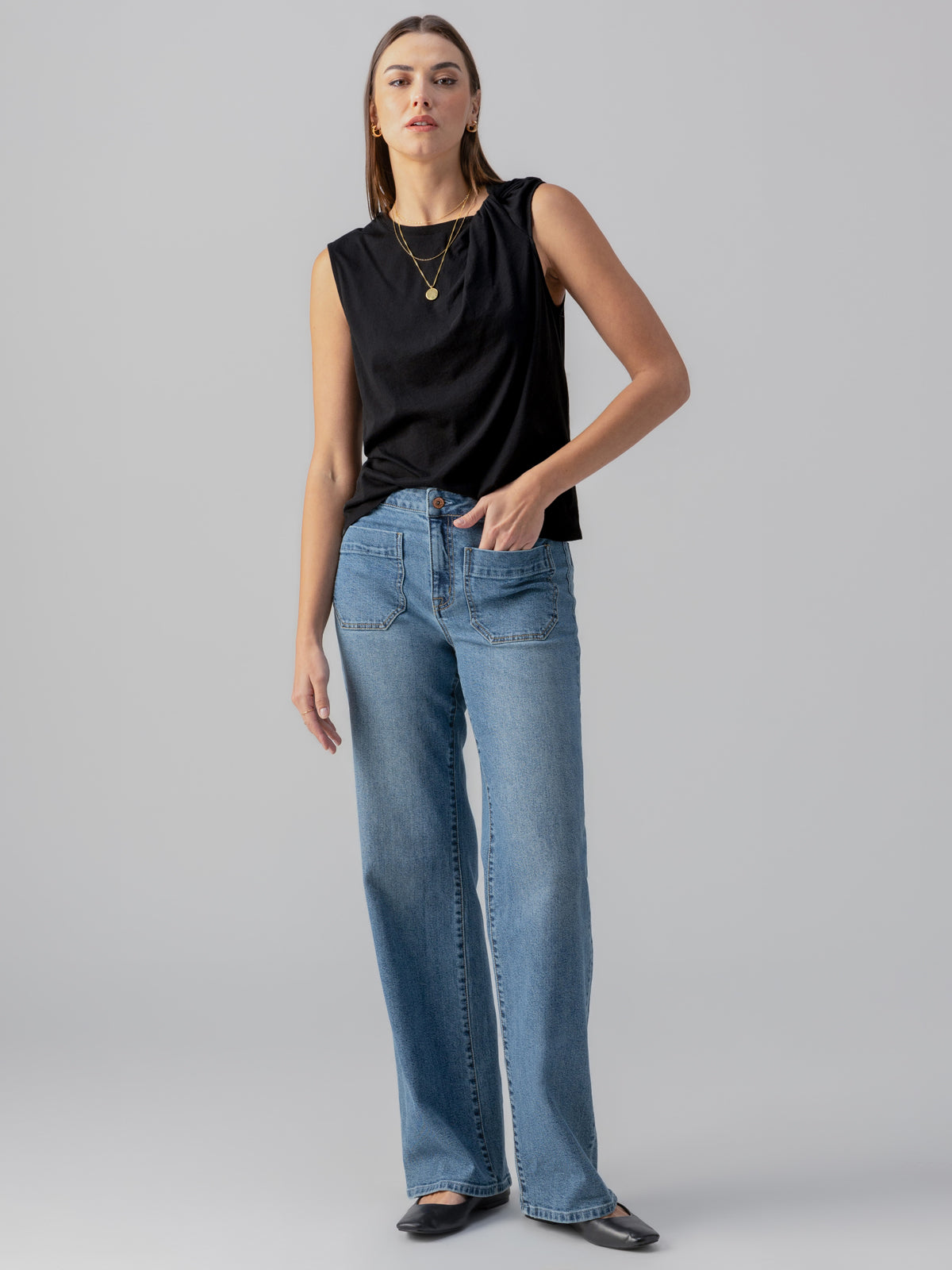 A woman stands against a plain gray background, wearing a sleeveless black top, the Marine Extended Pant in Sapphire Dream from Sanctuary Clothing, and black shoes. She has her right hand in her pocket and is wearing a gold layered necklace. She gazes ahead with a neutral expression.