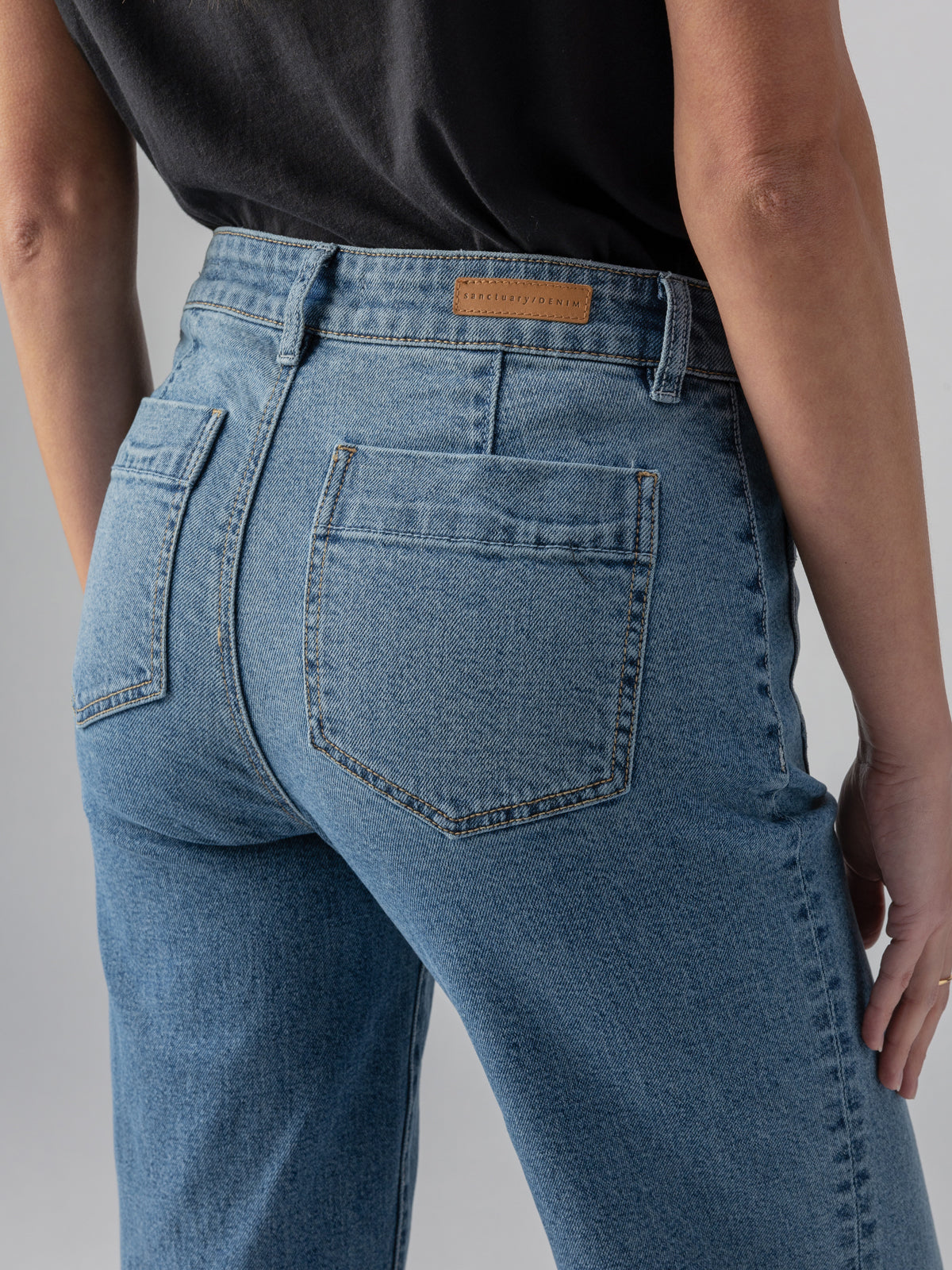 A person wearing a black top is shown from the back, highlighting their light blue Marine Extended Pant Sapphire Dream jeans from Sanctuary Clothing. The jeans feature two back pockets and a leather patch on the waistband. The background is plain and neutral.