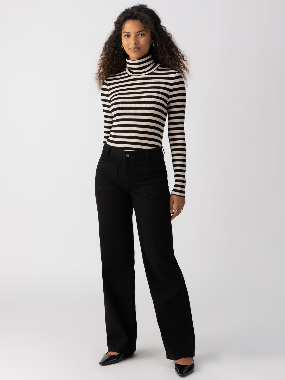 A person is standing against a plain background, wearing the Marine Full Length Black sweater from Sanctuary Clothing, along with black wide-leg pants. They have long, curly hair and are wearing black pointed shoes. They are looking forward with a neutral expression.