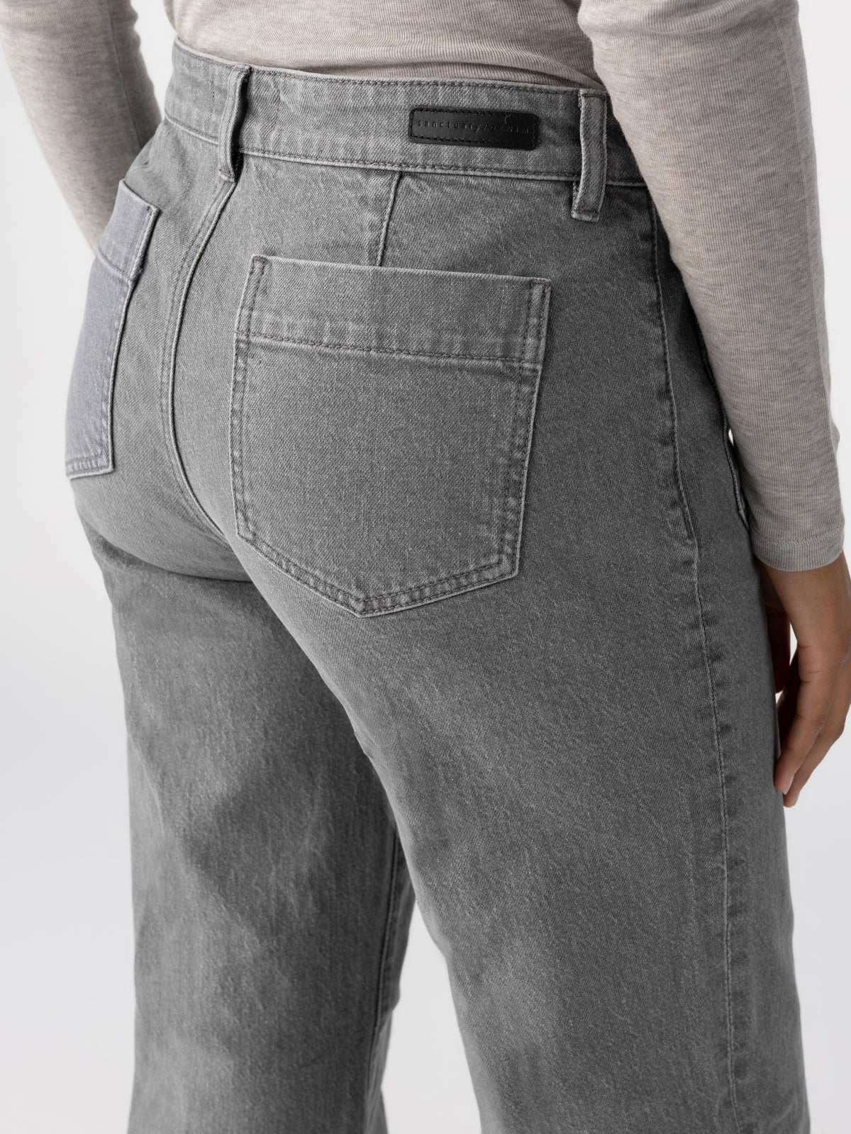 A person is wearing the Marine Full Length Asphalt high-waisted jeans by Sanctuary Clothing and a light gray long-sleeved shirt. The image focuses on the back side, highlighting the fit and style of the jeans, including the back pockets and belt loops.