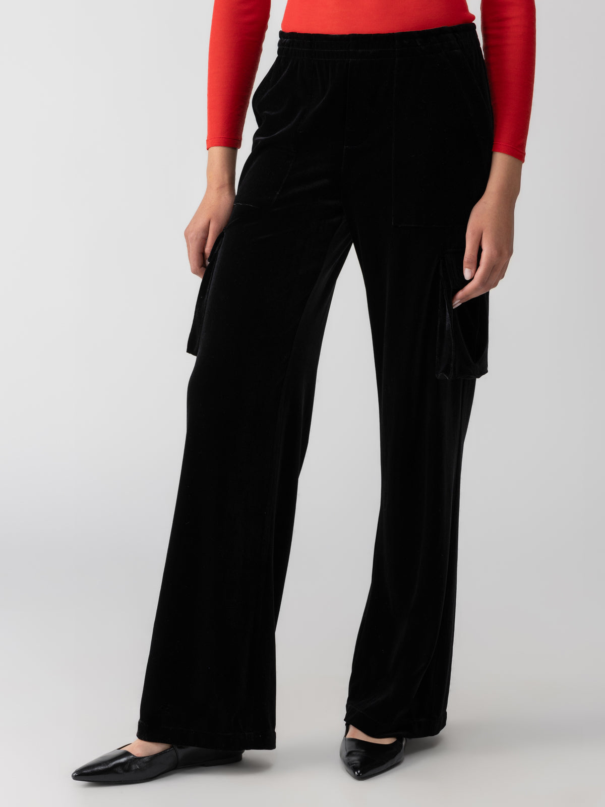 A person is seen from the shoulders down, wearing a bright red long-sleeve top and loose-fitting Sanctuary Clothing's Relaxed Velvet Reissue Black pants with side pockets. They have black pointed-toe flats on their feet. The background is plain white.