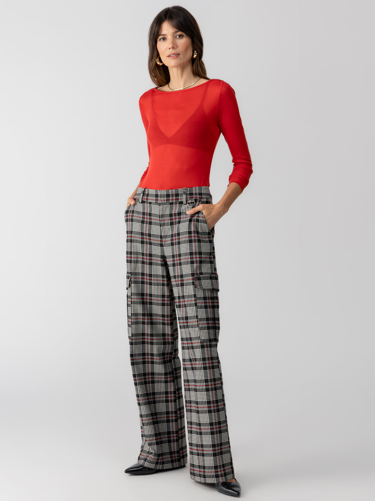 A woman stands against a plain backdrop, wearing a long-sleeve, semi-sheer red top and high-waisted, wide-leg "Gwen Cargo Garnet Plaid" trousers from Sanctuary Clothing with pockets. She has her hands in her pockets and is looking directly at the camera. She completes the outfit with black pointed-toe shoes.