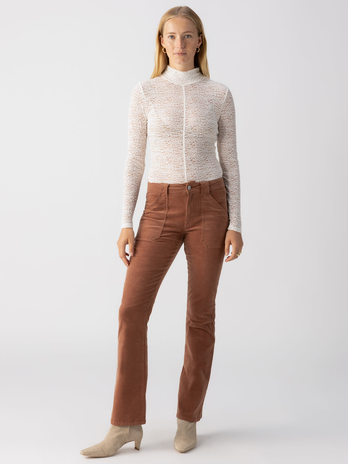 A woman with long blonde hair is standing against a plain background. She is wearing a sheer, long-sleeved white top paired with the Corduroy Hayden Bootcut Raw Hide pants from Sanctuary Clothing, which are brown and high-waisted with front pockets. She completes her look with beige ankle boots and minimal jewelry, including hoop earrings.