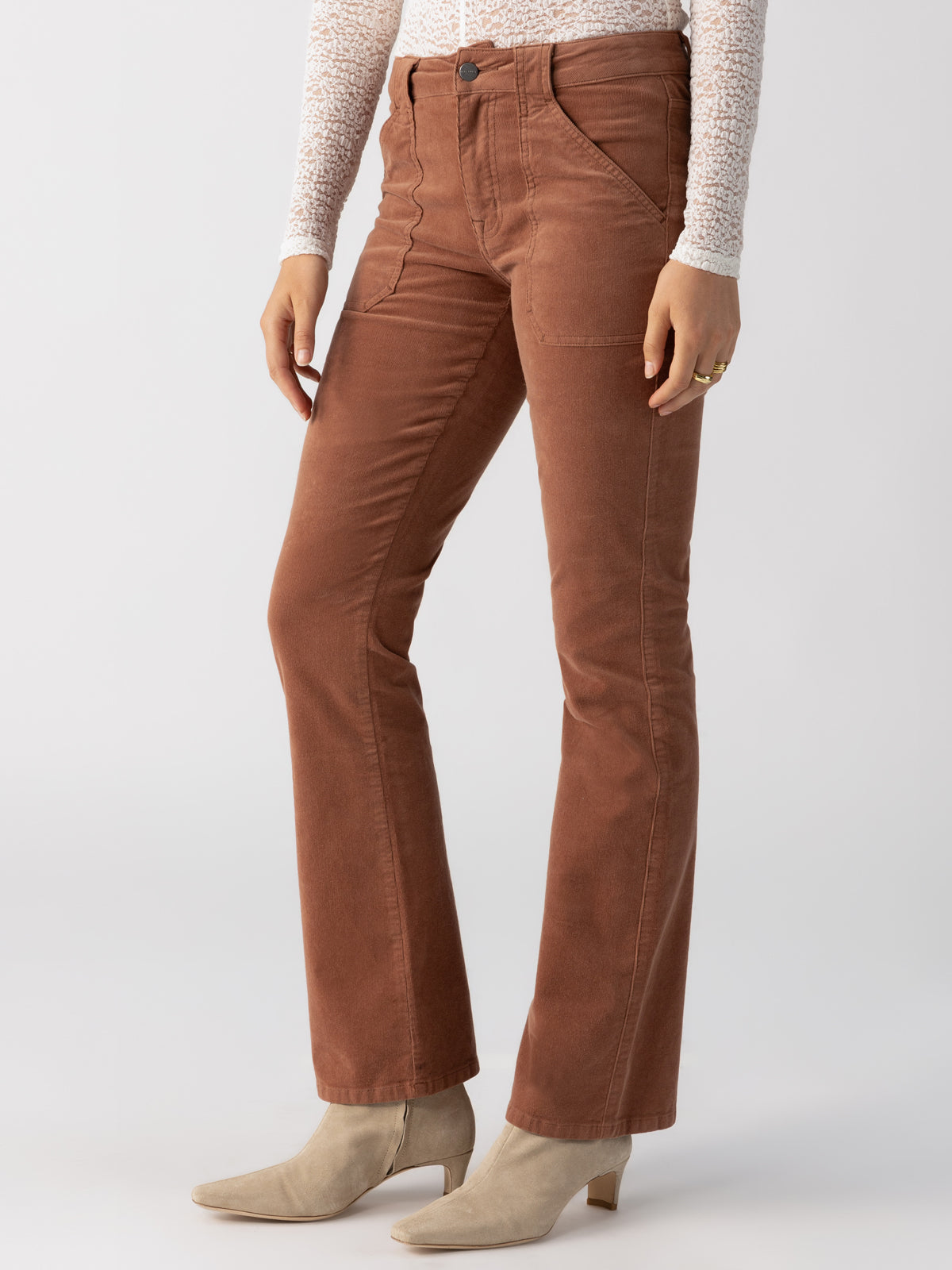 A person wearing Sanctuary Clothing's Corduroy Hayden Bootcut Raw Hide trousers in brown and a white long-sleeve top. They are also wearing light beige ankle boots. The photo is cropped from the shoulders down and is set against a plain white background.