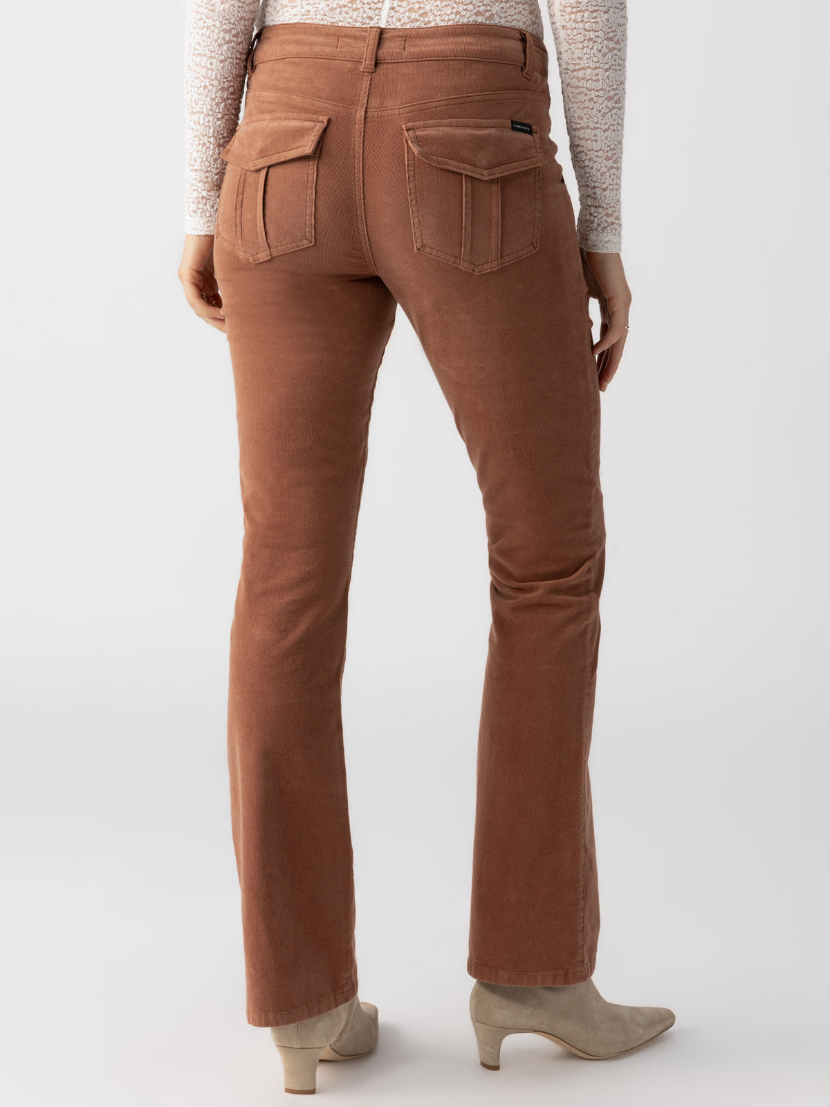 A person is shown from the back, wearing Sanctuary Clothing's Corduroy Hayden Bootcut Raw Hide pants with patch pockets and flared legs, paired with beige ankle boots. The individual is also wearing a light, long-sleeved top. The background is plain white.