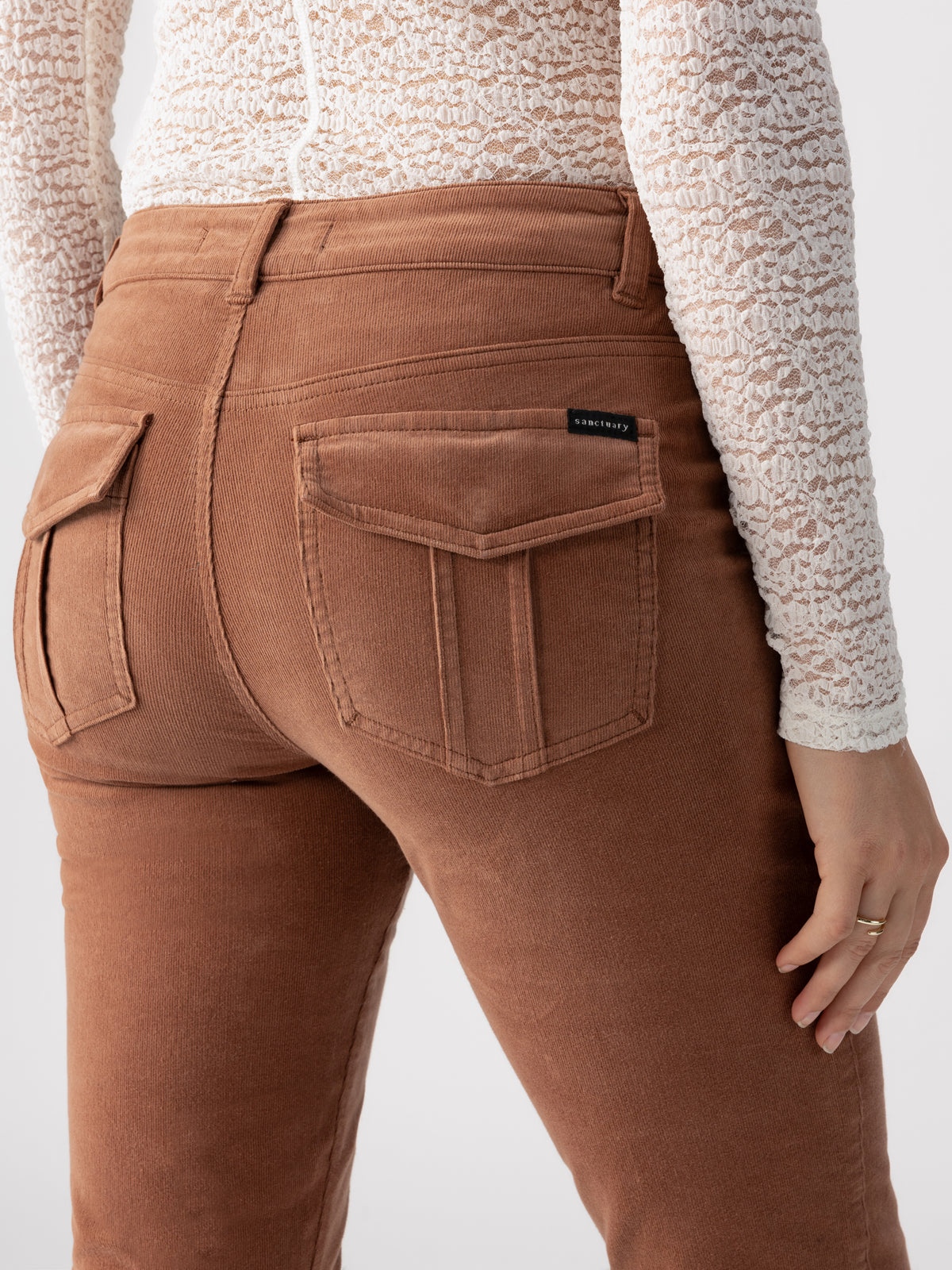 A person is shown from the back wearing Sanctuary Clothing's Corduroy Hayden Bootcut Raw Hide pants, which are high-waisted with large back pockets. They are also wearing a white, long-sleeve lace top. The person’s right hand, adorned with a ring, is resting near their right pocket.