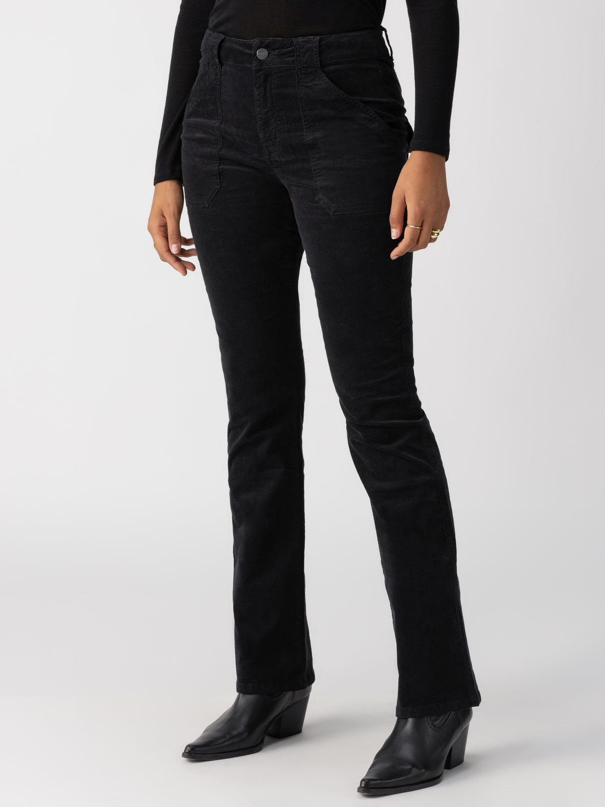 A person wearing the Velveteen Hayden Bootcut Pant in Black by Sanctuary Clothing and a black long-sleeve top is standing against a plain white background. They have their arms relaxed by their sides and are wearing black pointed-toe boots. Only the lower body and part of the upper torso are visible.