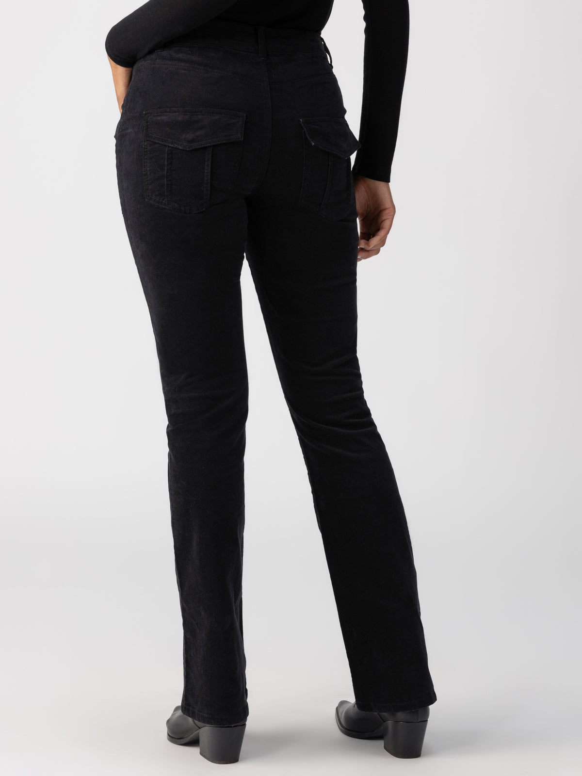 A person wearing the Velveteen Hayden Bootcut Pant in Black by Sanctuary Clothing, which features both front and back pockets, is shown from the back. They are standing in a studio setting against a plain background, with their lower half visible. They are also wearing black heeled boots and a black long-sleeve top.