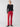 A person with light hair stands confidently against a plain background, wearing a long-sleeve black top, the Velveteen Hayden Bootcut Pant in Mars Red by Sanctuary Clothing, and black shoes. They have one hand in a pocket and an expressive look on their face.