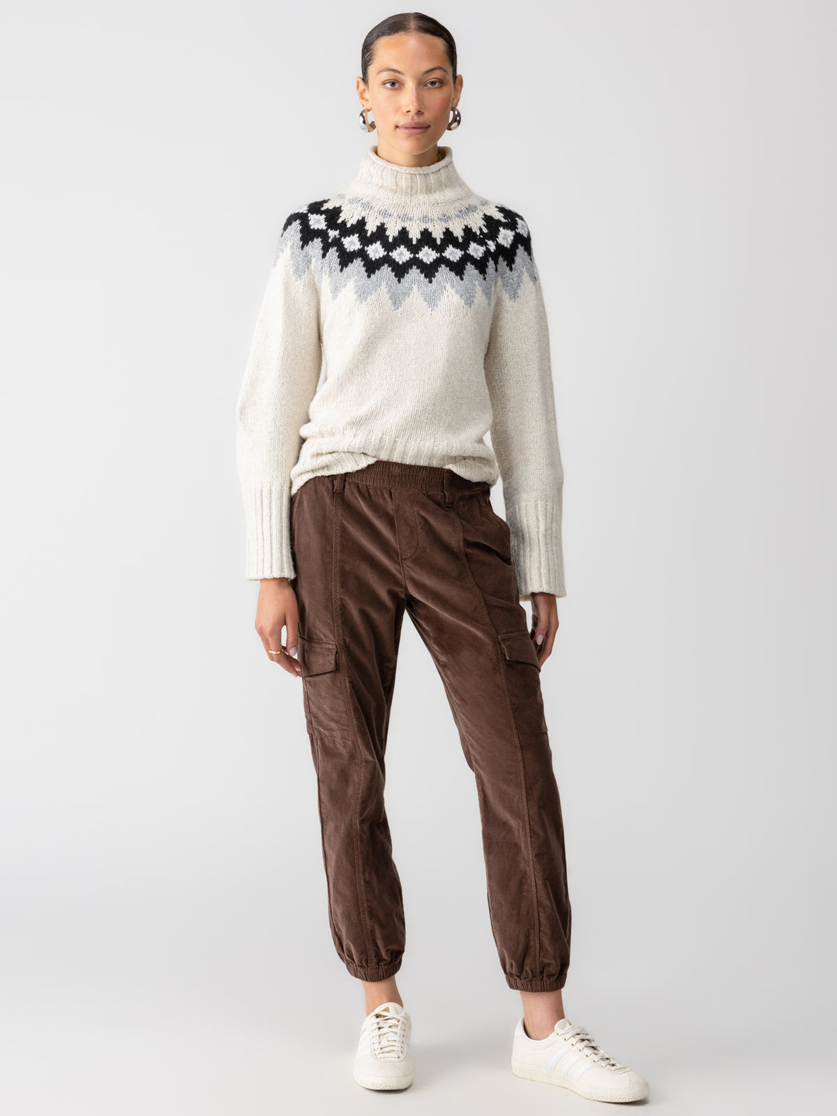 A person stands against a plain background wearing a beige turtleneck sweater with a black and white zigzag pattern across the chest and brown cargo pants from Sanctuary Clothing, named "Corduroy Relaxed Rebel Brown Sugar." They have white sneakers on and gold hoop earrings, posing with their hands by their sides.