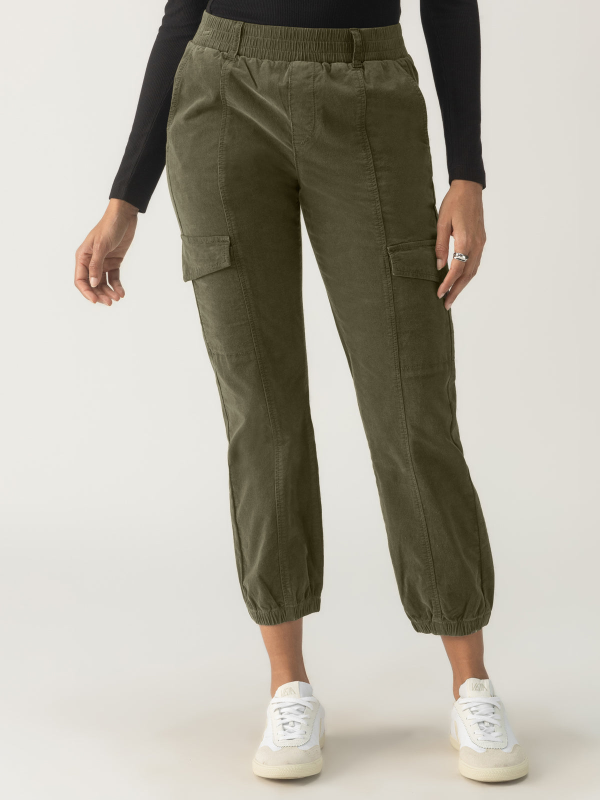 A person wearing Sanctuary Clothing's Corduroy Relaxed Rebel pants in Burnt Olive stands against a plain background. They are also wearing a black long-sleeve top and white sneakers. One hand is casually placed in a pocket while the other hand rests by their side.