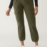 A person wearing Sanctuary Clothing's Corduroy Relaxed Rebel pants in Burnt Olive stands against a plain background. They are also wearing a black long-sleeve top and white sneakers. One hand is casually placed in a pocket while the other hand rests by their side.