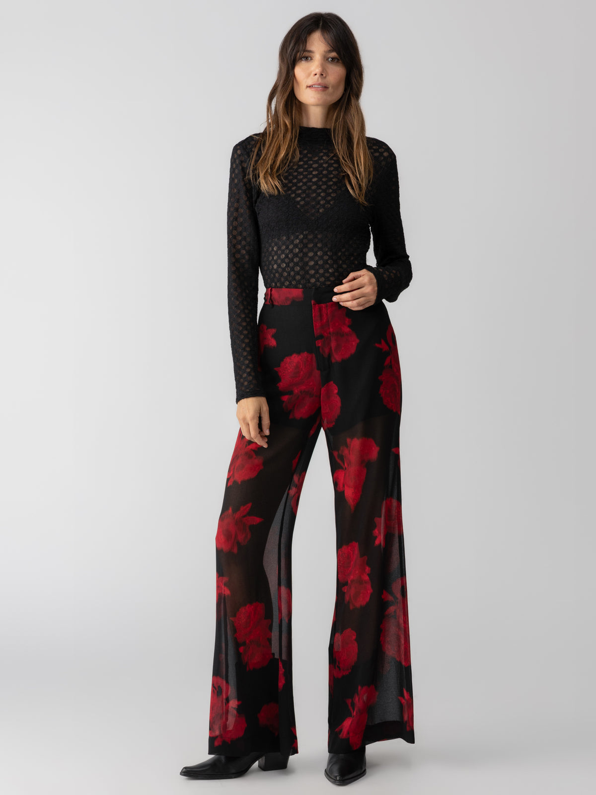 A person with long brown hair is wearing a sheer black top with a circular pattern and a pair of high-waisted, wide-leg "Sheer Nights Pant Moody Rose" from Sanctuary Clothing, adorned with large red floral prints. They are posing against a plain gray background.