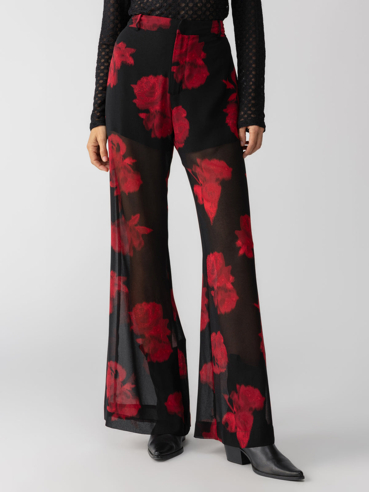 A person is wearing the Sheer Nights Pant Moody Rose by Sanctuary Clothing, featuring a bold red floral pattern on black sheer wide-leg trousers. They paired this striking look with pointed black boots and a dark textured long-sleeved top. The background is plain white.