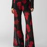 A person is wearing the Sheer Nights Pant Moody Rose by Sanctuary Clothing, featuring a bold red floral pattern on black sheer wide-leg trousers. They paired this striking look with pointed black boots and a dark textured long-sleeved top. The background is plain white.