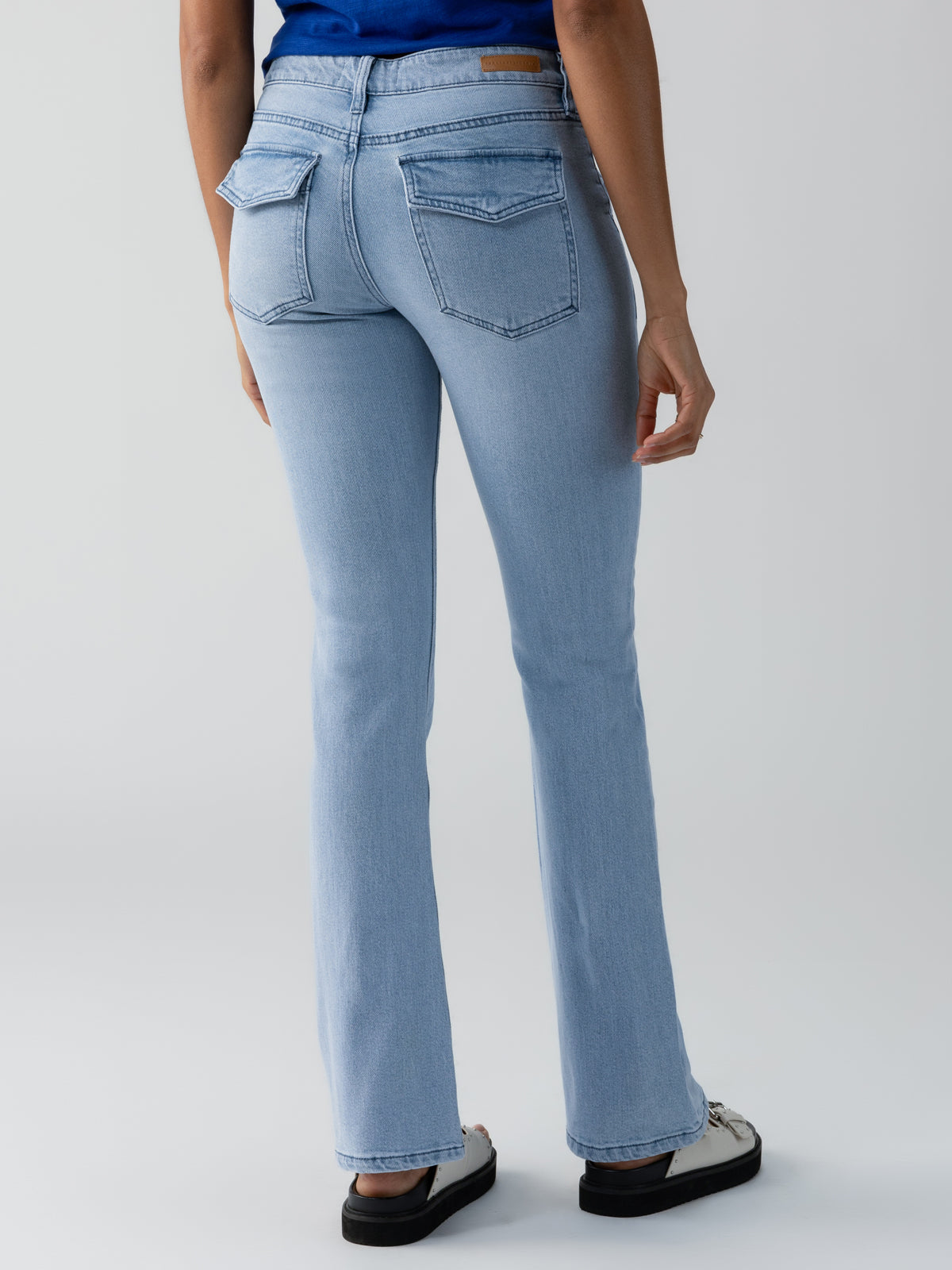 A person wearing The Revival Bootcut Jean Bliss by Sanctuary Clothing and a blue top is shown from the back. The jeans have two back pockets with flaps and flare slightly at the bottom. The person is also wearing white sneakers with black soles. The background is a plain light grey.