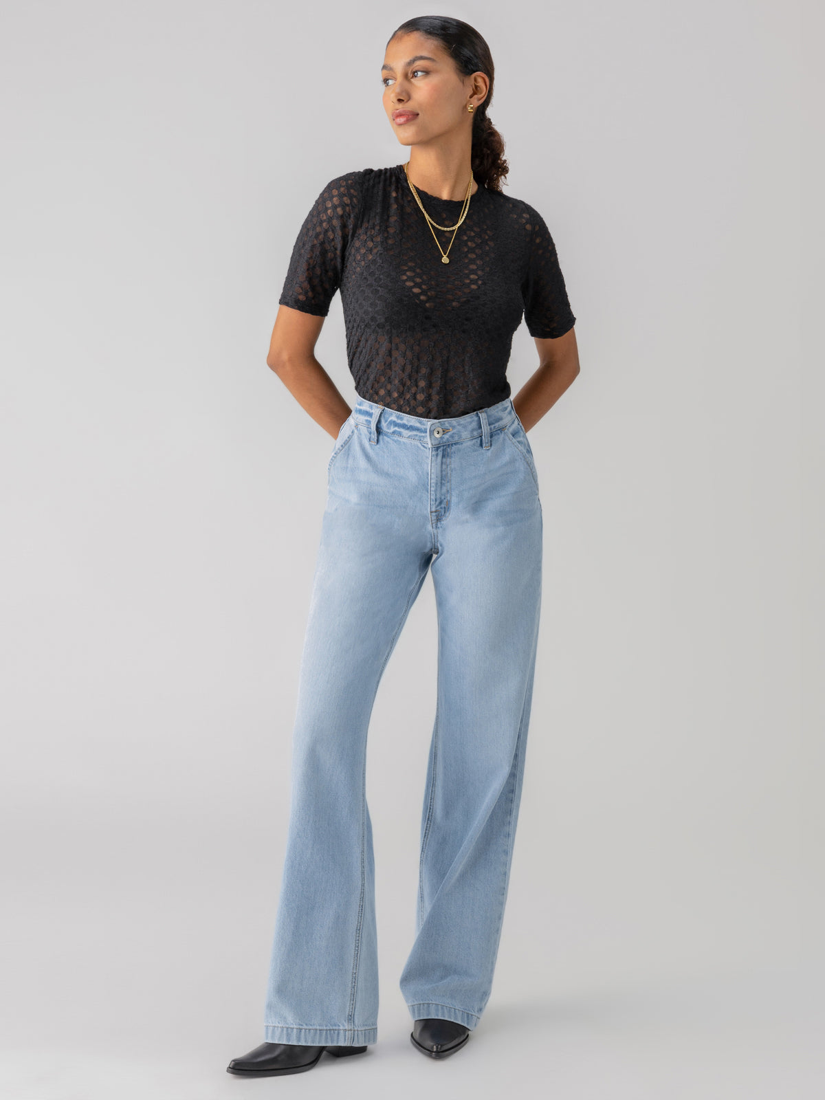 A person stands against a plain background wearing a black, short-sleeved, sheer top with a dotted pattern, tucked into the Tailored Renewal Jean Mirage from Sanctuary Clothing. They have their hands in their pockets and are accessorized with layered gold necklaces and small hoop earrings.