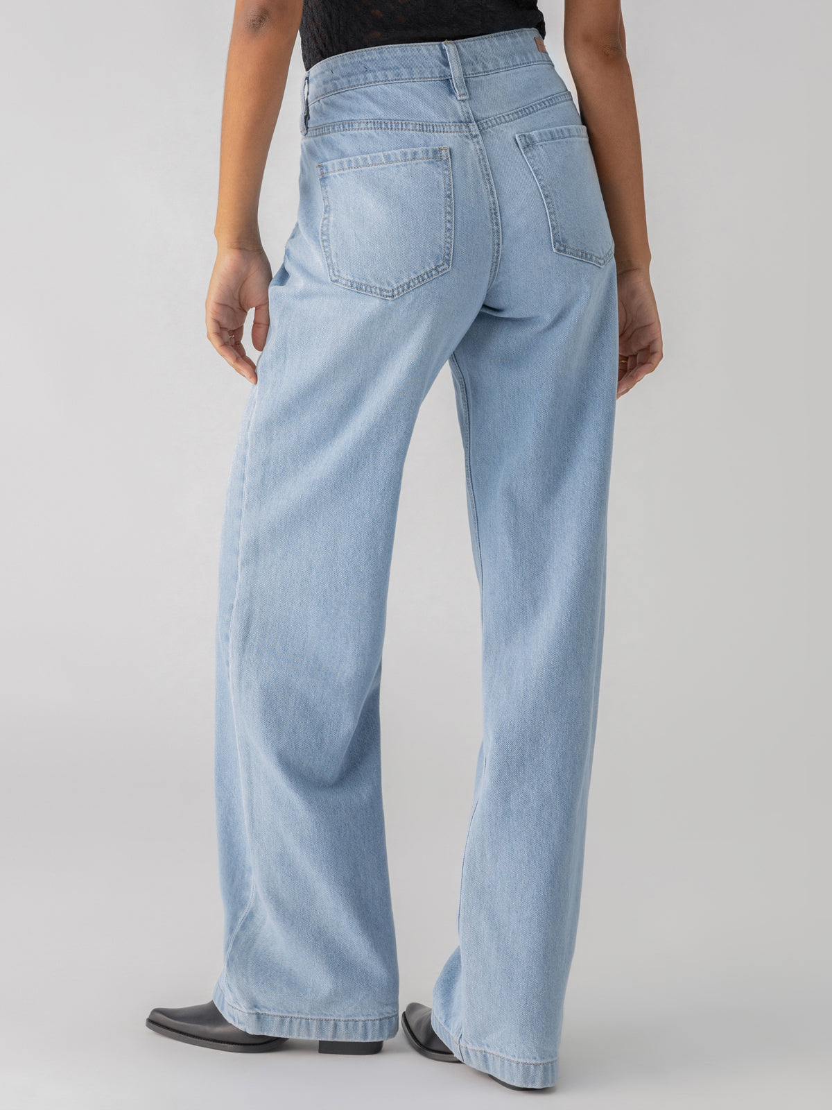 A person wearing the Tailored Renewal Jean Mirage from Sanctuary Clothing stands with their back facing the camera. The light blue wide-leg jeans have two back pockets and are paired with a black top. The person is standing against a plain, light-colored background.