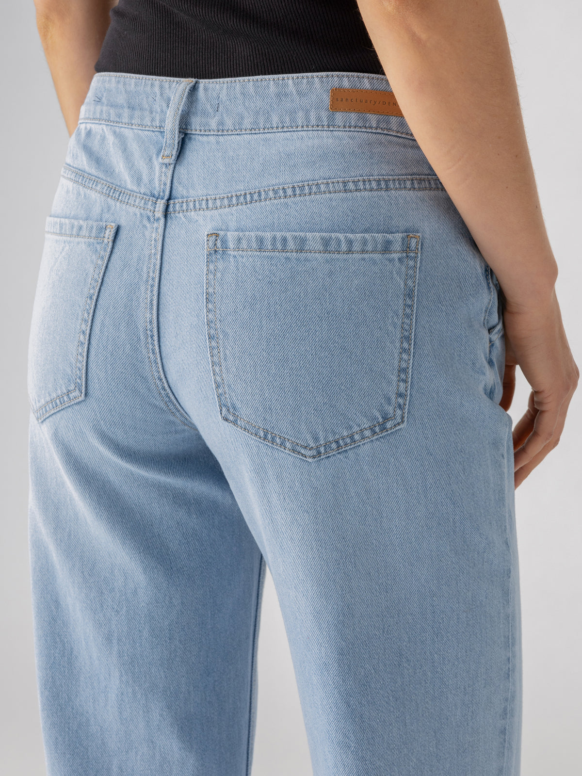 Close-up of a person wearing the Tailored Renewal Jean Mirage by Sanctuary Clothing, shown from the back. The light blue denim jeans feature two back pockets and light stitching. The person is also clad in a dark top, with their right hand partially visible.