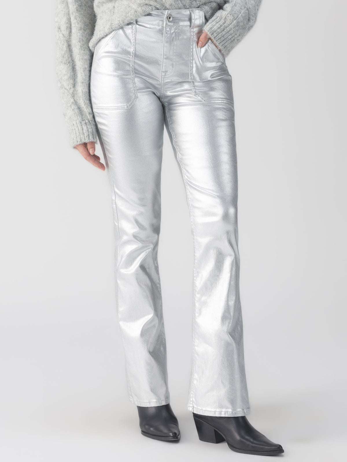 An individual dressed in the Sanctuary Clothing Metallic Hayden Bootcut Pant in Silver paired with a textured gray sweater and black heeled boots stands against a plain background.