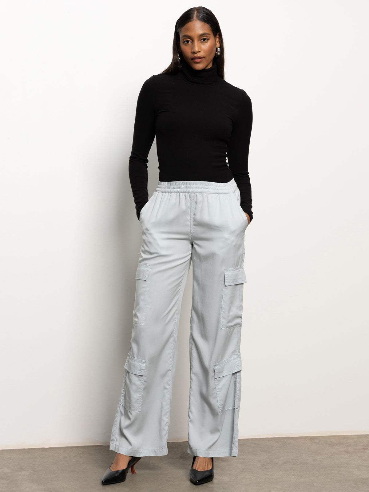 A person stands against a plain background, dressed in a black long-sleeve turtleneck and the Sanctuary Clothing "down to earth soft cargo" pants in pearl blue. They have their hands in their pockets and are wearing black heels.