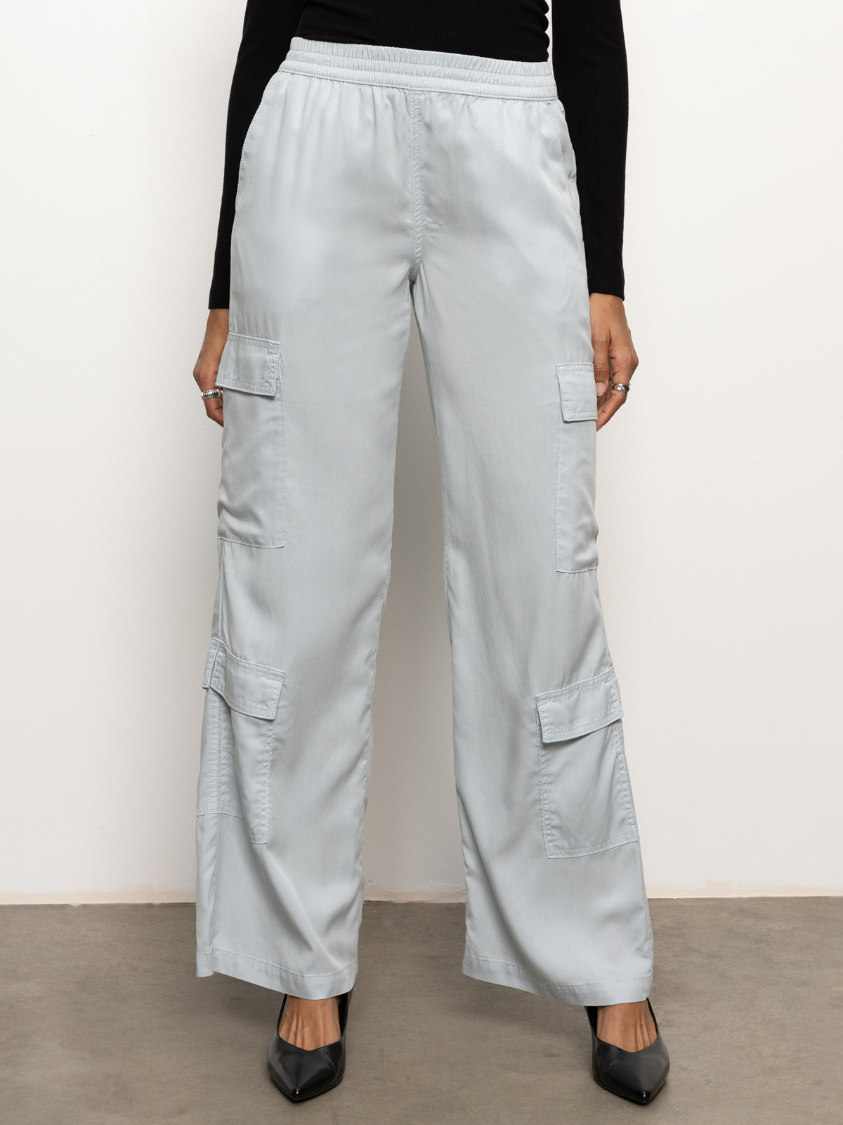 A person wearing Sanctuary Clothing's "down to earth soft cargo" pants in pearl blue, featuring multiple pockets, stands against a plain white background. They have paired these cargo pants with a long-sleeve black top and black pointed shoes. Only the lower half of the body is visible.